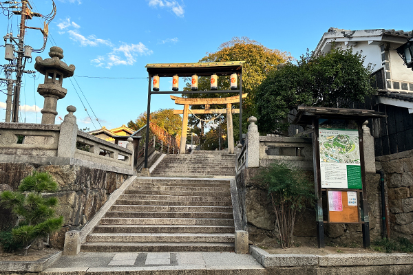 倉敷美観地区の北東にある阿智神社の参道入り口。青空の下、歴史を感じさせる長い石段と石造りの鳥居、そして夜には灯りがともる献灯台が並ぶ、街の守護神としての神聖な景観